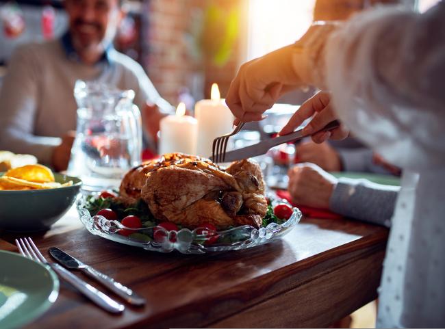 A person carves a turkey at Thanksgiving dinner. Several people can be seen sitting around the table. The table is set with festive plates and candles.