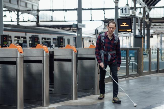 A man using a white cane leaving a railway station. In the background are the ticket gates and a stationary commuter train.